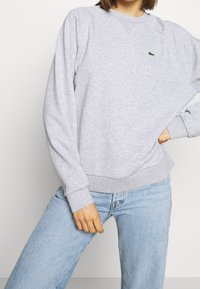 Sudadera gris claro con un cuello redondo acanalado, mangas largas y un pequeño logo verde en el pecho. Combinada con jeans azul claros.