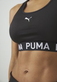 Crna sportska brusa s dizajnom leđa na racer, koja ima bijeli Puma logo i debelu crnu traku s natpisom "PUMA" u bijelim slovima. Glatka tekstura.