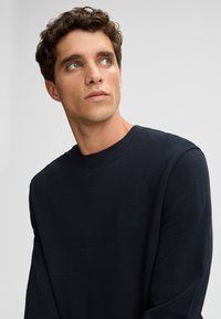 Chemise à manches longues bleu marine avec un col rond et un tissu texturé. Le modèle regarde légèrement vers le haut contre un fond neutre.