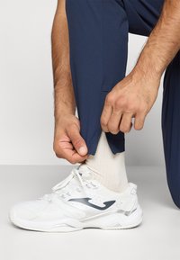 Λευκά αθλητικά παπούτσια με γκρίζες λεπτομέρειες, σε συνδυασμό με ναυτικά παντελόνια που έχουν φερμουάρ στον αστράγαλο. Η υφή φαίνεται λεία.