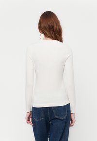 Donna con lunghi capelli rossi, che indossa un top aderente bianco a maniche lunghe e jeans blu, in piedi contro uno sfondo bianco semplice, di spalle.