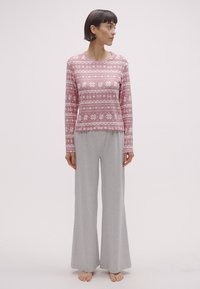 Różowa bluzka z długim rękawem z białymi kwiatowymi wzorami, zestawiona z szerokimi jasnoszarymi spodniami z miękkiego materiału. Modelka stoi boso na jednolitym tle.