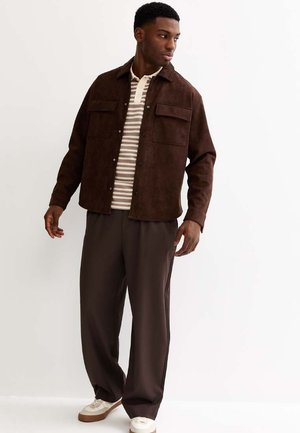 Uomo con giacca in camoscio marrone scuro, polo a righe beige e bianca, pantaloni larghi scuri e scarpe da ginnastica bianche, in piedi e che guarda di lato.