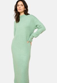 Svetlo zelena dolga pletena obleka z visokim ovratnikom. Ima sproščen kroj in stranske žepe. Mehka tekstura in dolžina do tal.