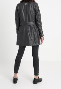 Manteau en cuir noir avec une taille ceinturée, des revers pointus et des poignets zippés, associé à un jean en denim foncé et des chaussures noires.
