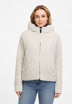 Frau trägt einen hellbeigen, gerippten Pullover mit Reißverschluss, Taschen und gepolstertem Kragen, kombiniert mit blauen Jeans, stehend vor weißem Hintergrund.