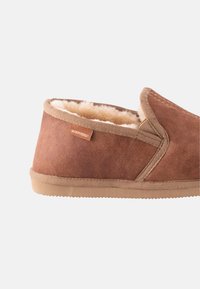 Brune ruskindssandaler med en blød beige pelsforing, rundet design, forstærkede syninger og en tan gummisål. Har et lille mærke.