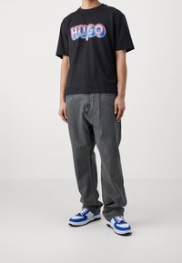 Svart bomullströja med färggrafik "HUGO", tillsammans med lösa grå denimjeans och blåvita sneakers.