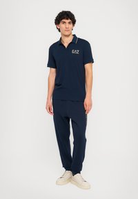 Mørkeblå polo-shirt med guld-logo og kontrastkrave, parret med matchende joggers med logo-print på siden og hvide sneakers.