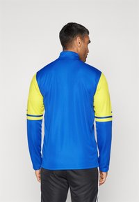 Blauwe en gele sportjack met lange mouwen en een ritskraag, met contrasterende mouwen en horizontale strepen op de armen.