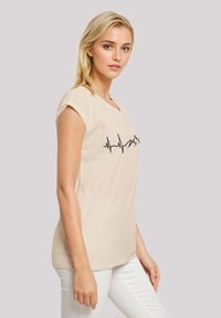 Beige cotton t-shirt with short sleeves, featuring a black heartbeat line graphic on the front. Fits loosely, with a rounded neckline.
