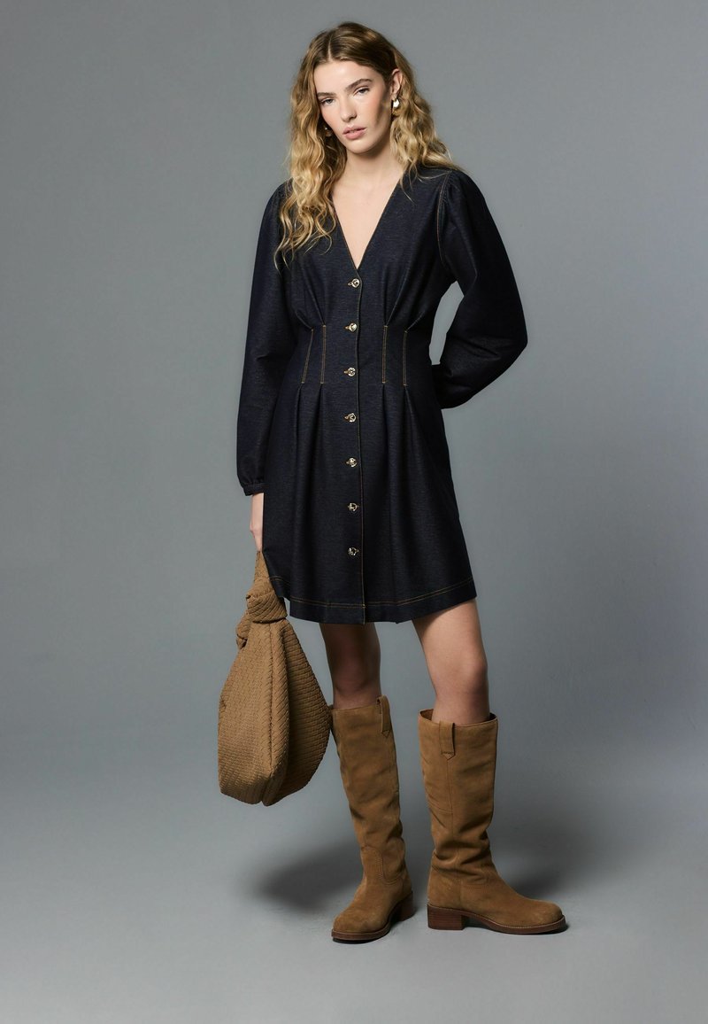 Džínové šaty tmavomodrej farby s dlhými nafúknutými rukávmi, hlbokým výstrihom do V, zapínaním na gombíky a zlatým prešívaním. Kombinované s hnedými čižmami po kolená a kabelkou.