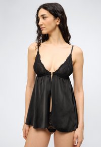 Frau trägt schwarze Spitze- und Satin-Unterwäsche mit dünnen Trägern, schaut nach links vor schlichtem hellem Hintergrund.