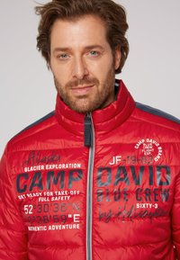 Giacca a vento rossa con collo alto, con testo e grafiche stampate in nero. Chiusura con zip e dettagli in contrasto blu navy sulle spalle.