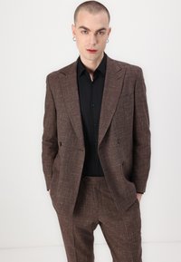 Costum în carouri maro cu sacou cu două rânduri de nasturi și pantaloni drepți, asortat cu o cămașă neagră cu nasturi. Textură netedă, croială ajustată.
