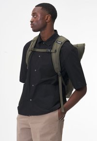 Olive-green, padded backpack with adjustable straps, worn over a black short-sleeve shirt. Features side pockets and a top handle.