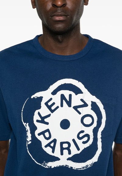 Mand iført en blå T-shirt med en stor hvid cirkulær logo med teksten "KENZO PARIS" på fronten.