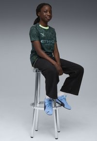 Sötétzöld póló geometrikus mintákkal, sárga kiemelésekkel és az "ETIHAD AIRWAYS" logóval. Fekete, bő szárú nadrággal és világoskék sportcipővel viselve.