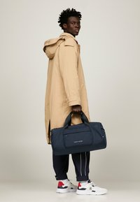Cappotto impermeabile beige, borsa duffel blu navy con tasca frontale, manici neri, sneakers bianche con accenti rossi e blu navy, e pantaloni della tuta blu navy.