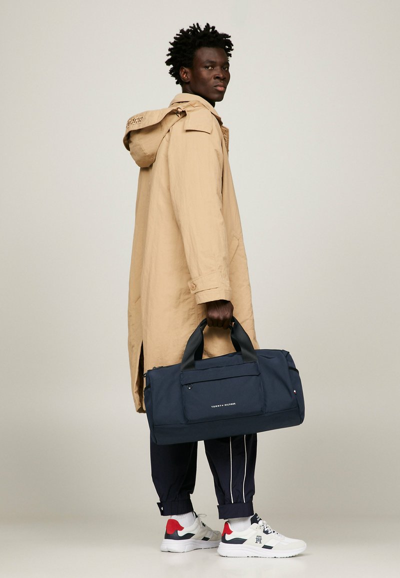 Cappotto impermeabile beige, borsa duffel blu navy con tasca frontale, manici neri, sneakers bianche con accenti rossi e blu navy, e pantaloni della tuta blu navy.