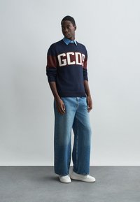 Tumed sviiter valgete ja burgundia "GCDS" logoga, kantud helesinise kraega särgi peal, koos laia jala helesiniste teksade ja valgete tossudega.