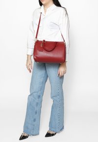 Borsa a tracolla rossa in pelle con finitura texture, manici doppi, tracolla regolabile e un'etichetta, abbinata a jeans a zampa d'elefante blu chiaro e tacchi neri.