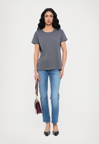 Grijze T-shirt met korte mouwen en een ronde hals, gecombineerd met lichtblauwe rechte jeans. Model houdt een bordeauxrode handtas vast en draagt zwarte hakken.