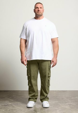 Hombre de pie mirando hacia adelante, vistiendo una camiseta blanca lisa, pantalones cargo verde oliva y zapatillas blancas sobre un fondo neutro.