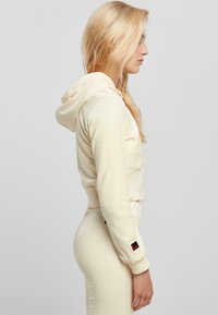 Hoodie crème, coupé, à la texture douce, avec capuche et poignets élastiques ; accent de logo sur la manche. Style ajusté visible de profil.