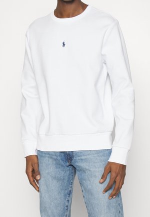 Mann trägt ein schlichtes weißes Sweatshirt mit langen Ärmeln und kleinem blauem Pferdelogo auf der Brust sowie hellblaue Jeans.