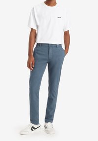 Vit T-shirt med rund hals och svart logotyp, kombinerad med blå bomullschinos och vita sneakers med svarta detaljer.