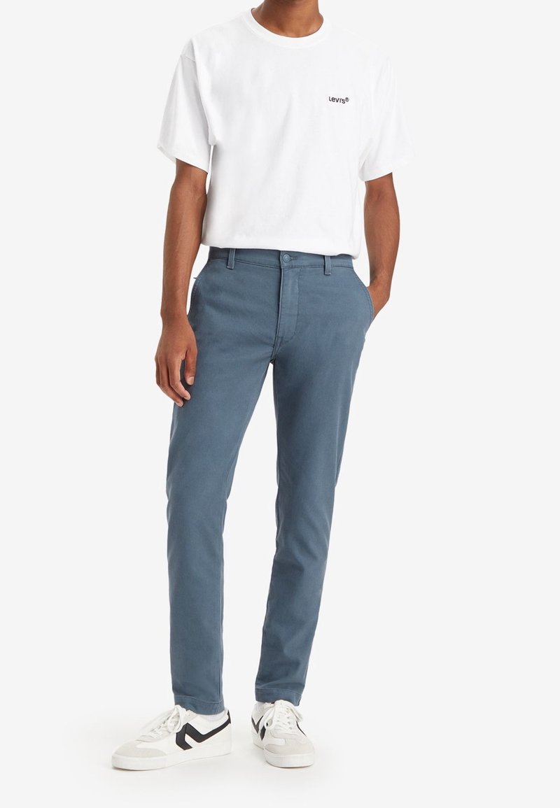 Vit T-shirt med rund hals och svart logotyp, kombinerad med blå bomullschinos och vita sneakers med svarta detaljer.