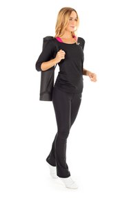 Black athletic top with three-quarter sleeves, paired with black leggings and white sneakers. A rolled yoga mat is slung over one shoulder.