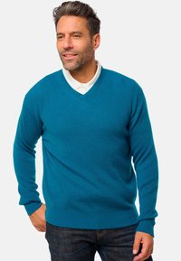 Pull en tricot côtelé teal avec un col en V, des manches longues et un ourlet ajusté. Porté par-dessus une chemise blanche à col avec un jean bleu.