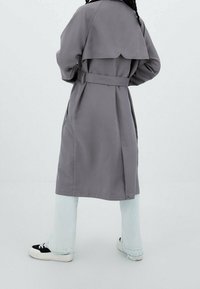 Grauer Trenchcoat mit breitem Kragen, tailliertem Gürtel und seitlichen Taschen. Verfügt über einen gefalteten Rücken und scheint aus einem glatten, leichten Stoff gefertigt zu sein.