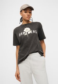 Νεαρό άτομο που φοράει ένα ξεθωριασμένο μαύρο μπλουζάκι με το γράμμα "Dreamers" και ένα λευκό γραφικό λουλούδι, ανοιχτό γκρι παντελόνι και μπεζ καπέλο μπέιζμπολ.