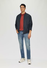 Veste bomber navy, polo rouge, jean bleu clair et baskets beiges. La veste présente une fermeture éclair et des poches latérales ; les chaussures ont des accents multicolores.