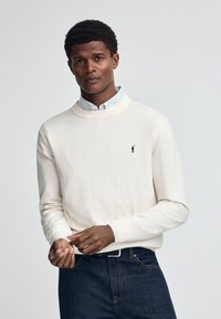 Cremfarbener Strickpullover mit Rundhalsausschnitt, versehen mit einem kleinen schwarzen Logo. Getragen über einem hellblauen gemusterten Hemd und kombiniert mit dunklen Jeans.