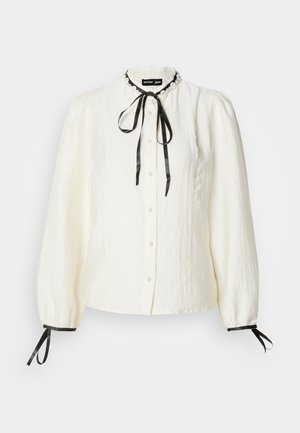 Fehér, hosszú ujjú blúz texturált anyagból, gombos elülső résszel, fekete szalaggal, és fekete díszítéssel a mandzsettán és a galléron. Puffos ujjakkal rendelkezik.