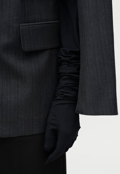 Mørkegrå blazer med subtile nålestriber, der har en opkrøllet ærme og sorte satin handsker, som fremhæver glatte teksturer og skræddersyet design.