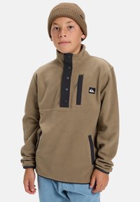 Pull en polaire marron avec patte de boutonnage noire et poche zippée. Dispose de poches latérales et d'un écusson logo. Bonnet en maille marron porté.