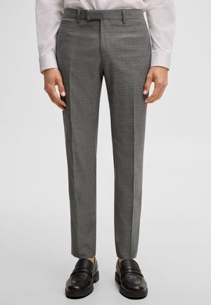 Mężczyzna w szarych wąskich spodniach garniturowych, białej koszuli i czarnych skórzanych mokasynach na jednolitym jasnym tle.