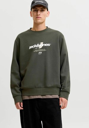 Nuori mies, jolla on päällään tummanvihreä Jack & Jones -collegepaita ja musta lippalakki, seisoo yksinkertaisen vaalean taustan edessä.