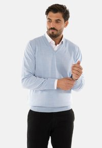 Maglione azzurro chiaro a V in tessuto knit leggero, con polsini e orlo a coste, indossato sopra una camicia bianca con colletto, abbinato a pantaloni neri.