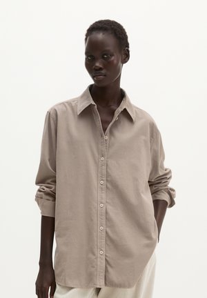 Personne grande à la peau foncée portant une chemise beige ample boutonnée aux manches retroussées et un pantalon clair sur un fond blanc uni.