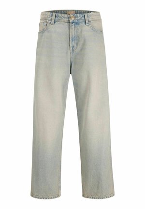 Uśmiechnięty chłopiec z krótkimi blond włosami, ubrany w prostą białą koszulkę, luźne jasnoniebieskie dżinsy i brązowe buty, stojący na jednolitym jasnym tle.