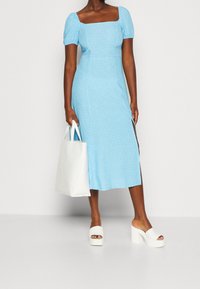 Jasnoniebieska sukienka z bufkami, kwadratowym dekoltem i kwiatowym wzorem. Białe sandały na obcasie oraz biała torba typu tote dopełniają stylizację.