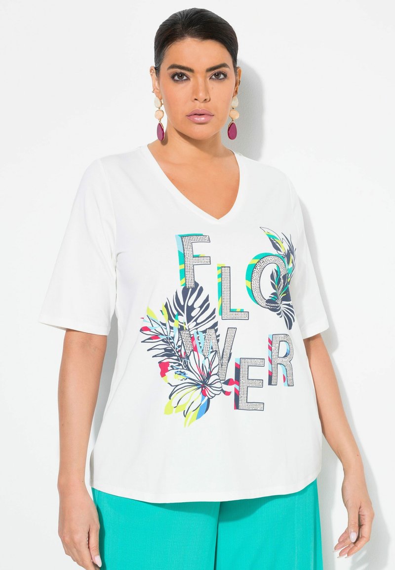 Camiseta blanca de manga corta con escote en V que presenta un colorido diseño floral y la palabra "FLOWER", adornada con lentejuelas. Combinada con pantalones en tono teal.