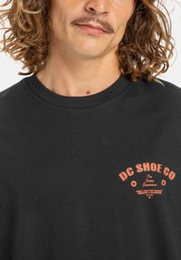 Zwarte katoenen t-shirt met een oranje grafische print "DC SHOE CO" op de borst, ronde hals en korte mouwen. Glad materiaal.