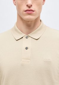 Νεαρός άνδρας που φοράει μπεζ μπλουζάκι πόλο BOSS με δύο κουμπιά κουμπωμένα, ορατός από το λαιμό μέχρι το μέσο του στήθους, με φόντο μια απλή επιφάνεια.